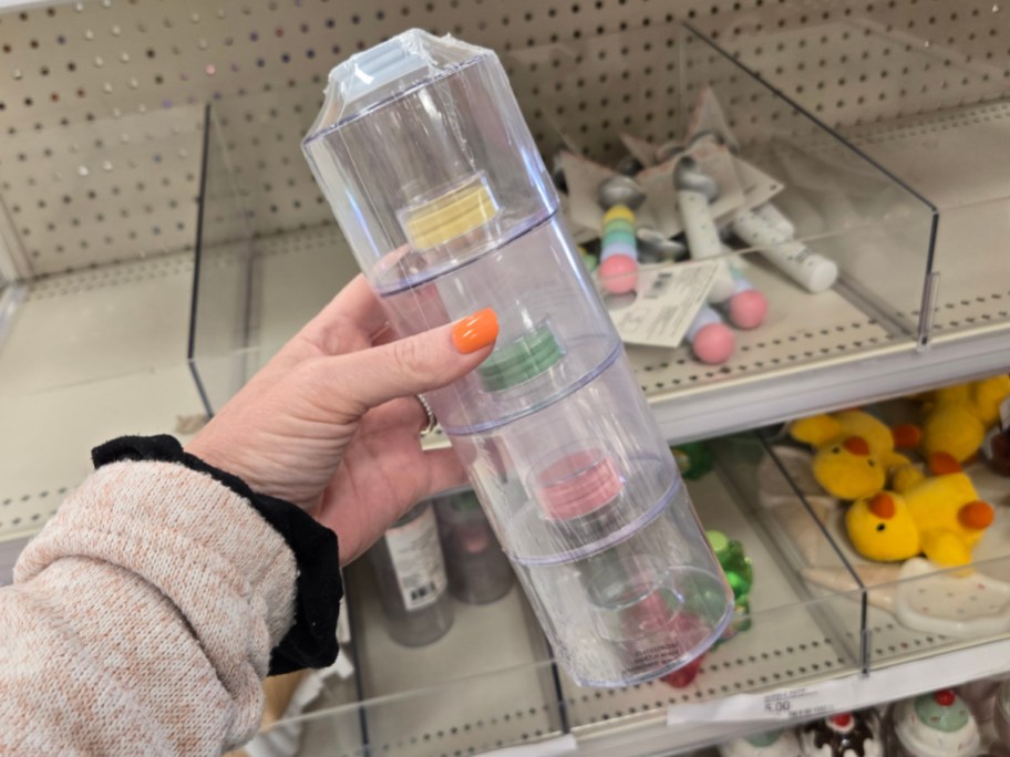 A hand with orange-painted nails holds a clear, stackable container with four lids in various pastel colors. Shelves in the background display assorted items.