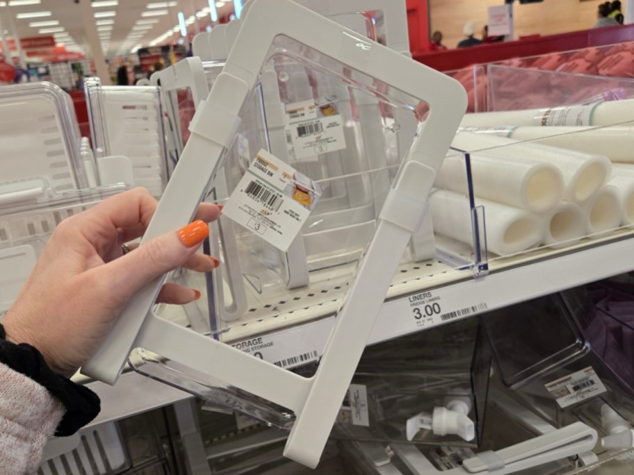 A hand with orange nails holds a white plastic storage organizer in a store aisle. Clear bins and rolled liners are visible in the background.