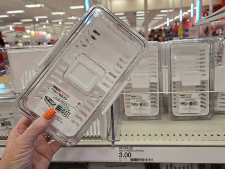 A hand with orange nail polish holds a transparent produce storage container. Several identical containers are displayed on a store shelf, priced at $3.00.