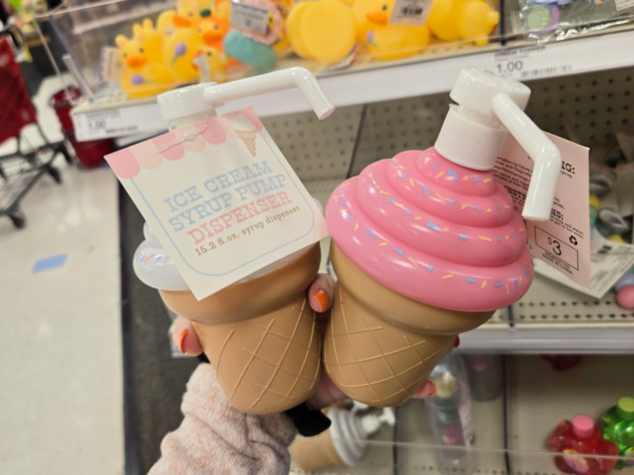 Two ice cream cone-shaped syrup pump dispensers with pink tops and sprinkles are held in a store. The tag reads