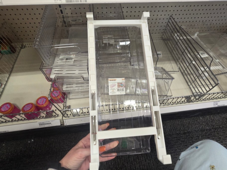 A hand holds a large, clear plastic container with a white frame in a store. Shelves in the background display similar containers and small colorful tubs.