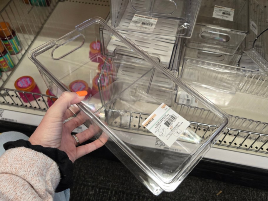 A hand holds a clear plastic storage container labeled with a price and barcode, in a store aisle with similar containers and colorful jars nearby.