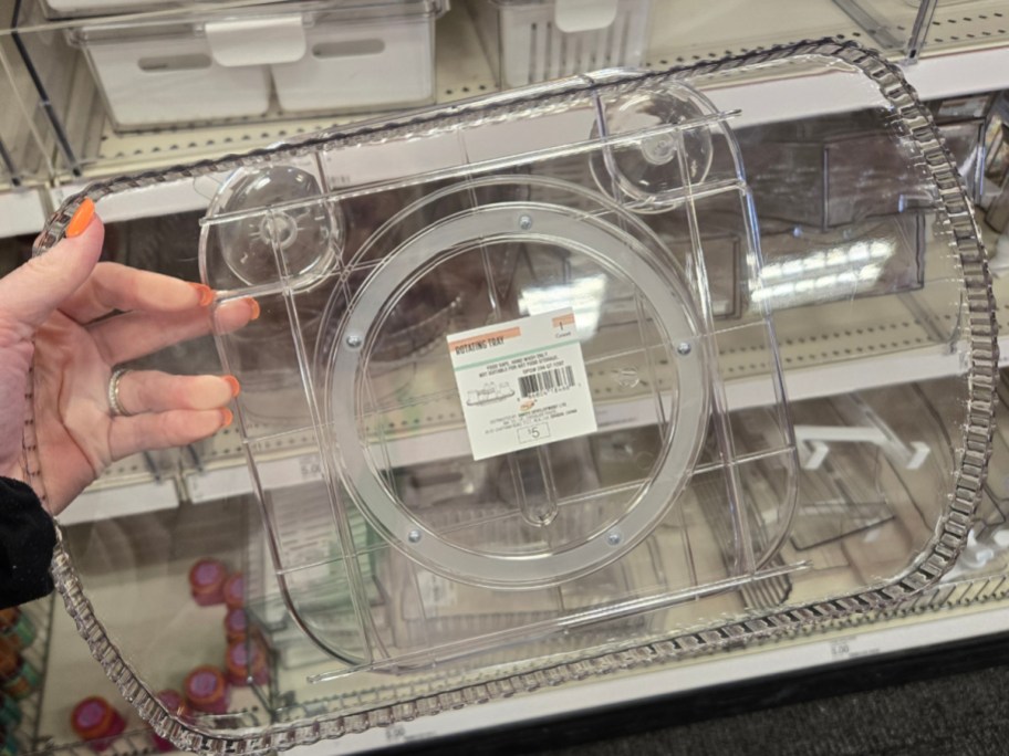 A person with orange nail polish holds a clear, rectangular storage container with multiple compartments. It's displayed on a store shelf.