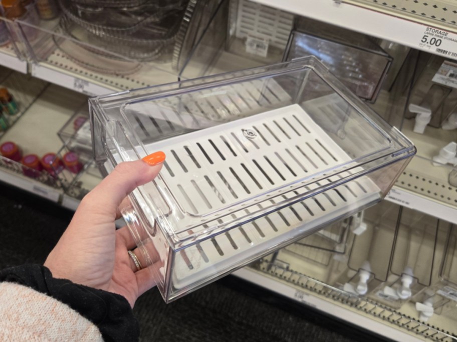A hand with an orange-painted nail holds a clear plastic storage container with a white slotted base, displayed in a store aisle with similar items.