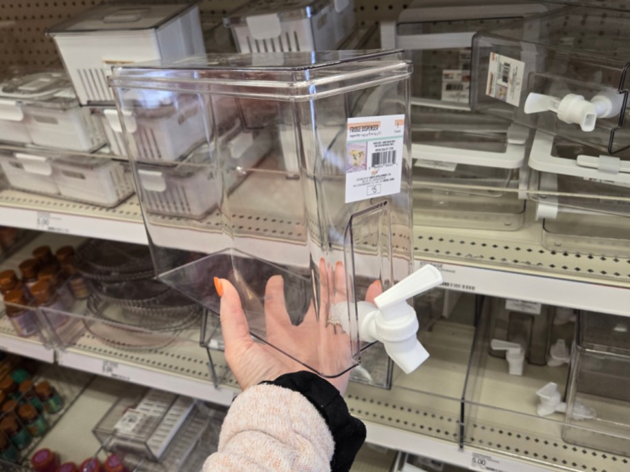 A hand holds a clear plastic beverage dispenser with a white spigot in a store aisle. Shelves display more dispensers and storage containers.