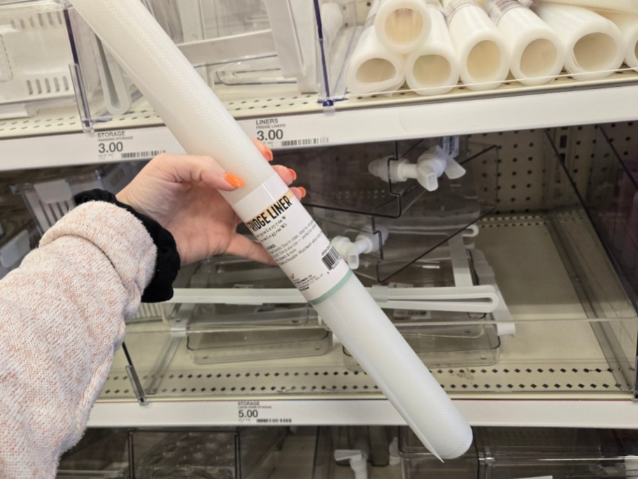 A person with bright orange nails holds a white drawer liner roll in a store aisle. Shelves display similar products, priced at $3.00 each.