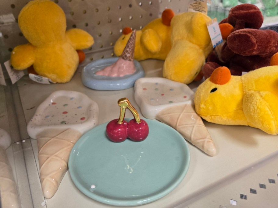 Stuffed yellow ducks and a brown bear sit beside ceramic dishes shaped like ice cream cones and cherries.