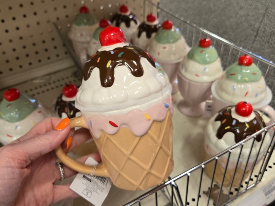 A hand with orange nail polish holds a mug shaped like an ice cream cone, with a whipped cream lid, chocolate drizzle, sprinkles, and a cherry. Numerous similar mugs are on a store shelf.