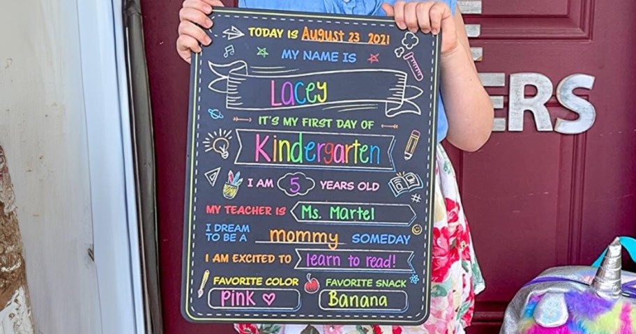 little girl standing outside her front door holding a first day of school chalkboard sign