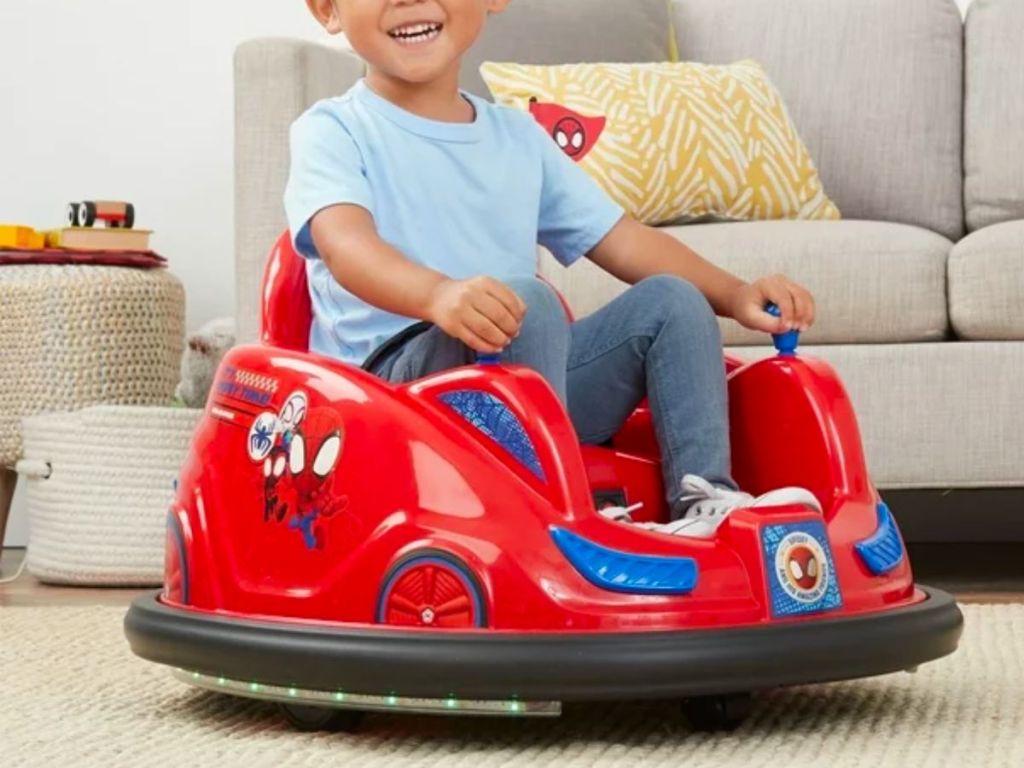 a little boy riding a Marvel's Spidey and His Amazing Friends 6V Bumper Car in a living room