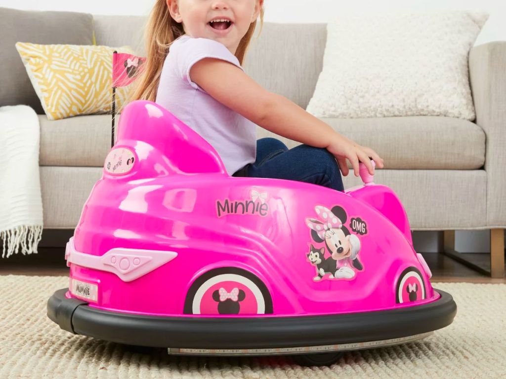a little girl riding a Disney's Minnie Mouse 6V Bumper Car in a living room