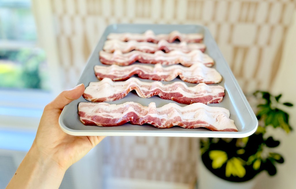 hand holding a sheet pan with raw bacon slices on top
