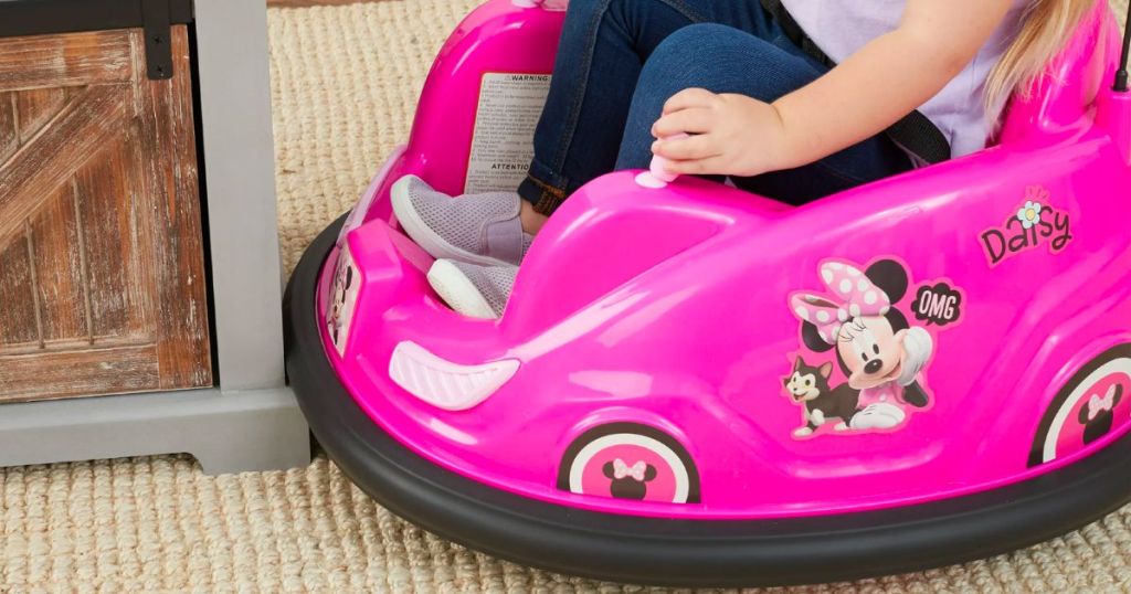 a little girl riding a Disney's Minnie Mouse 6V Bumper Car in a living room