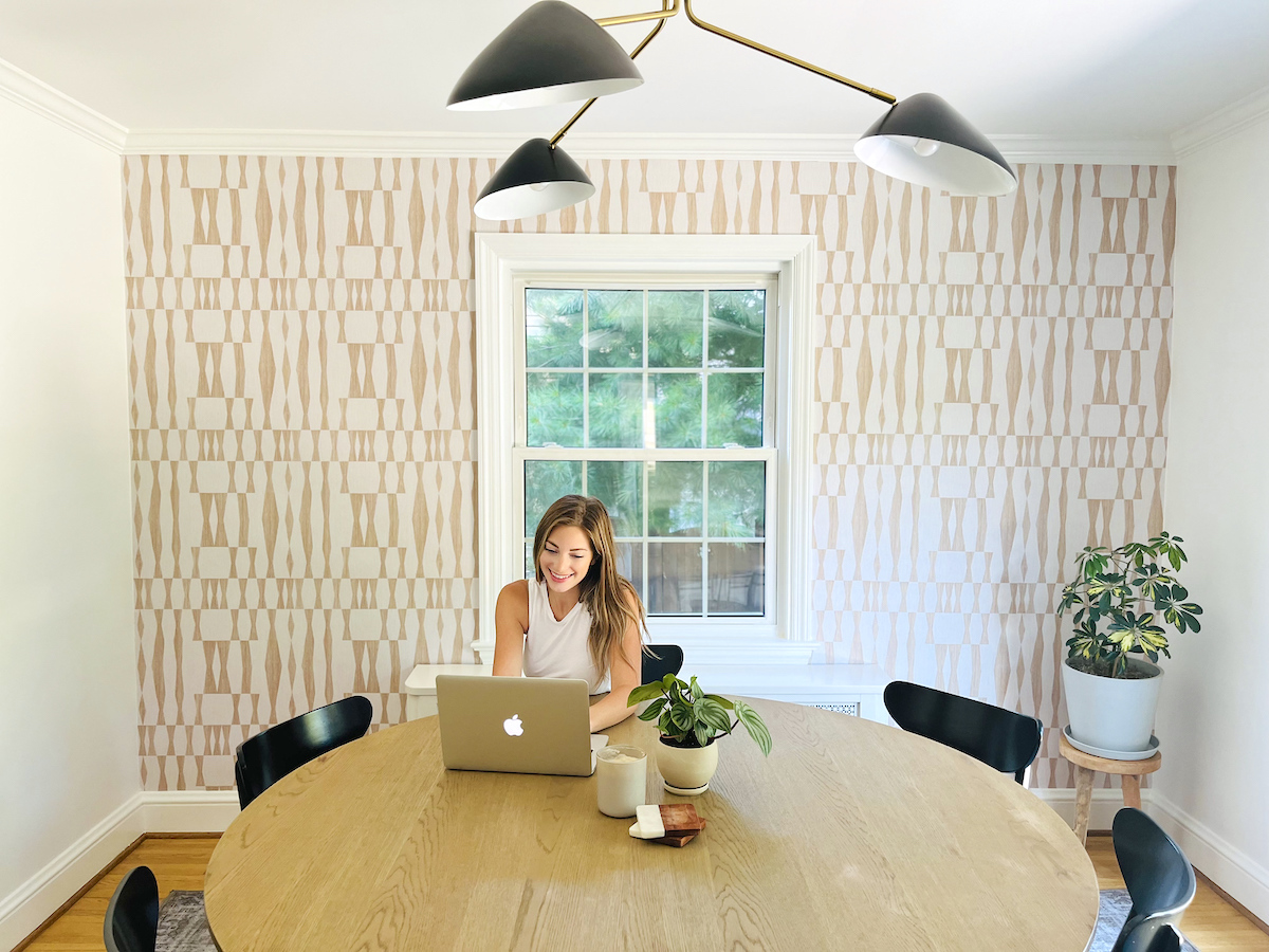 woman sitting at dining room table with apple laptop and patterned peel and stick wallpaper on wall in background