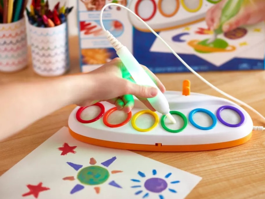 a kid's hand choosing a color on a toy coloring activity set, paper with drawings in front of the color chooser