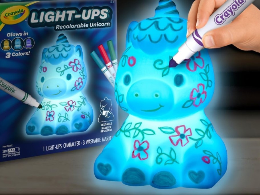 child's hand with a Crayola marker drawing on a a light up unicorn with doodles on it, box this toy comes in behind it