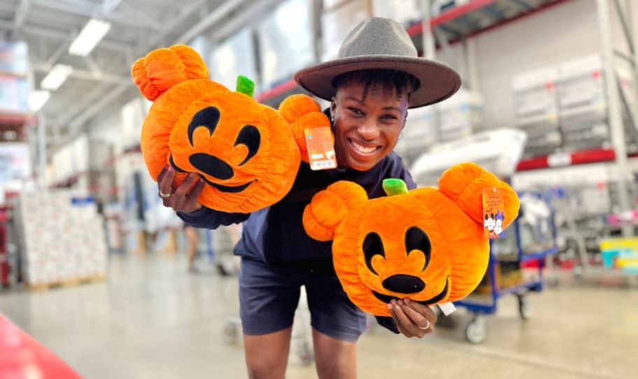 a woman in a hat holding a mickey mouse pumpkin plush in each hand