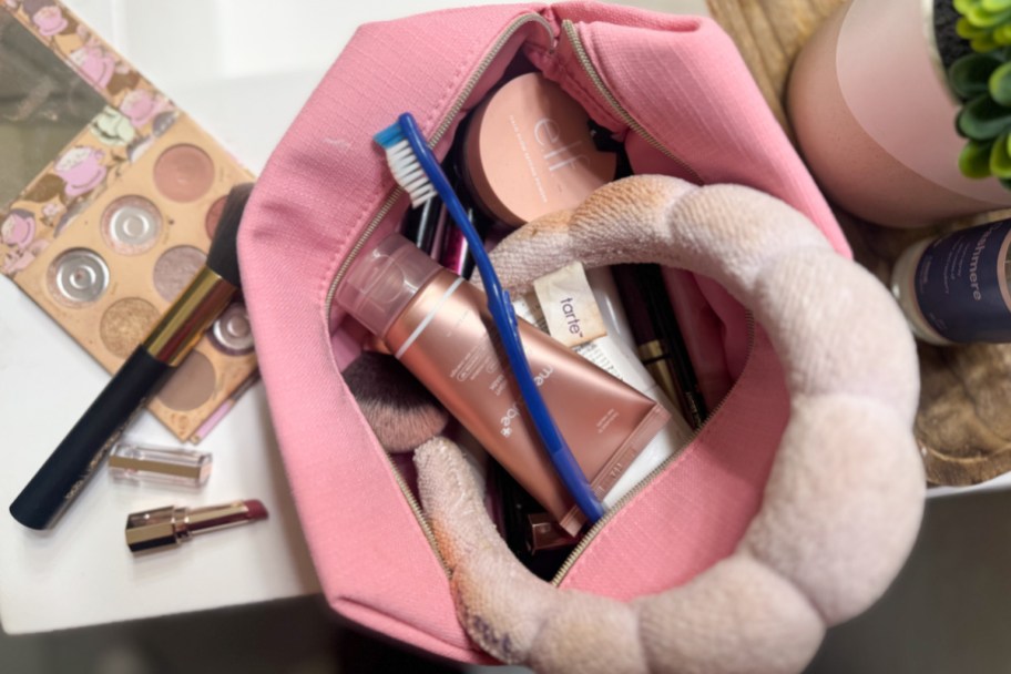 pink makeup bag opened with pink headband, toothbrush, makeup palette, and more makeup