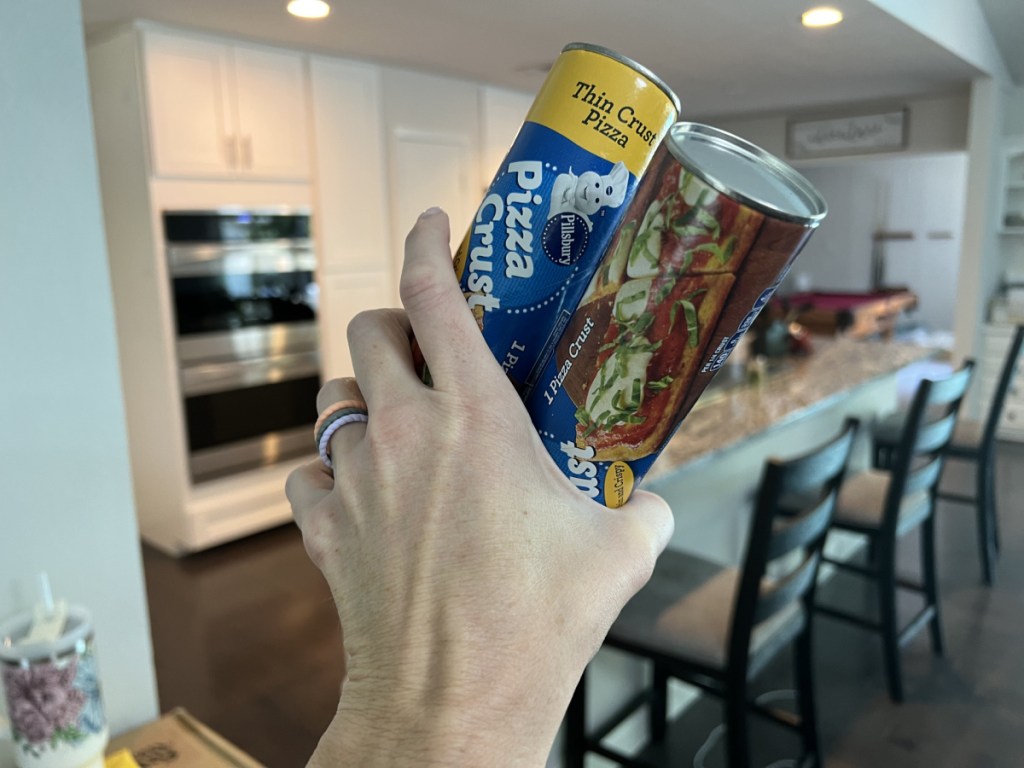 woman holding two cans of pillsbury pizza crust in front of a kitchen area