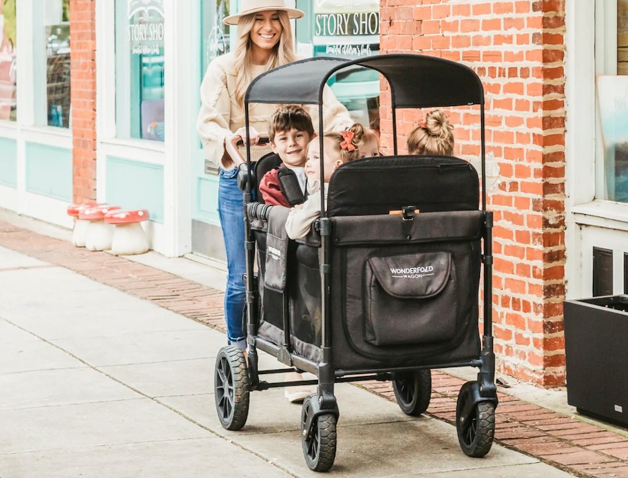 woman walking on sidewalk pushing wagon full of kids