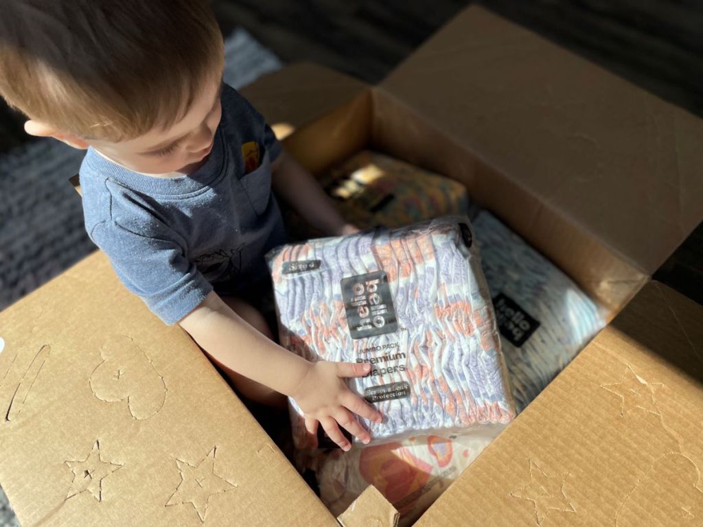 Baby sitting in a box holding a pack of Hello Bello diapers