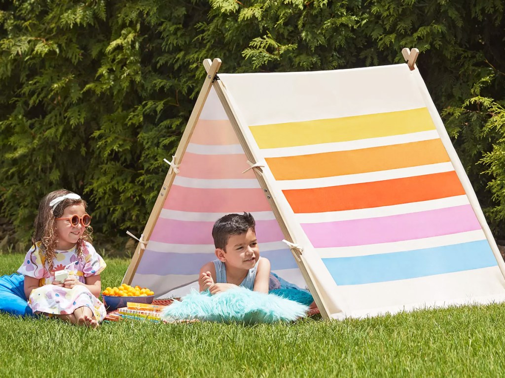 two kids sitting next to colorful striped play tent in grass
