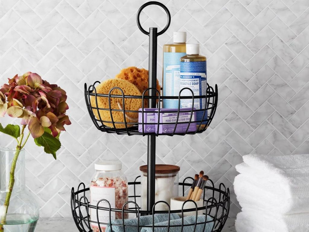 Two tier metal basket with bathroom supplies in it like washcloths, soap, bath salts, and folded towels next to it.
