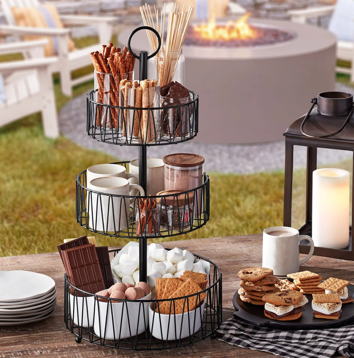 Three tiered basket holding supplies for s'mores like graham crackers, marshmallows, chocolate, and a row of hot chocolate supplies too.