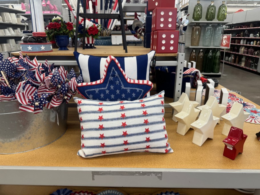Patriotic-themed store display featuring star-shaped pillows, small American flags in a metal bucket, and star decor. Colors are red, white, and blue.