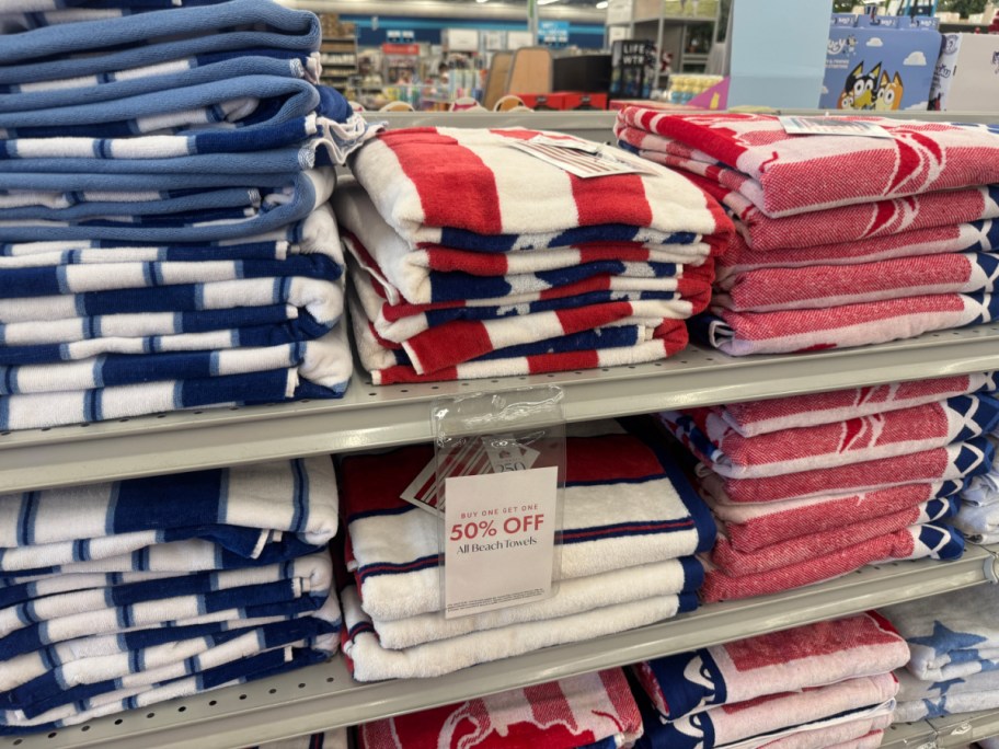 Stacks of colorful striped beach towels in red, white, and blue are neatly arranged on store shelves. A sign reads &ldquo;50% Off.&rdquo;