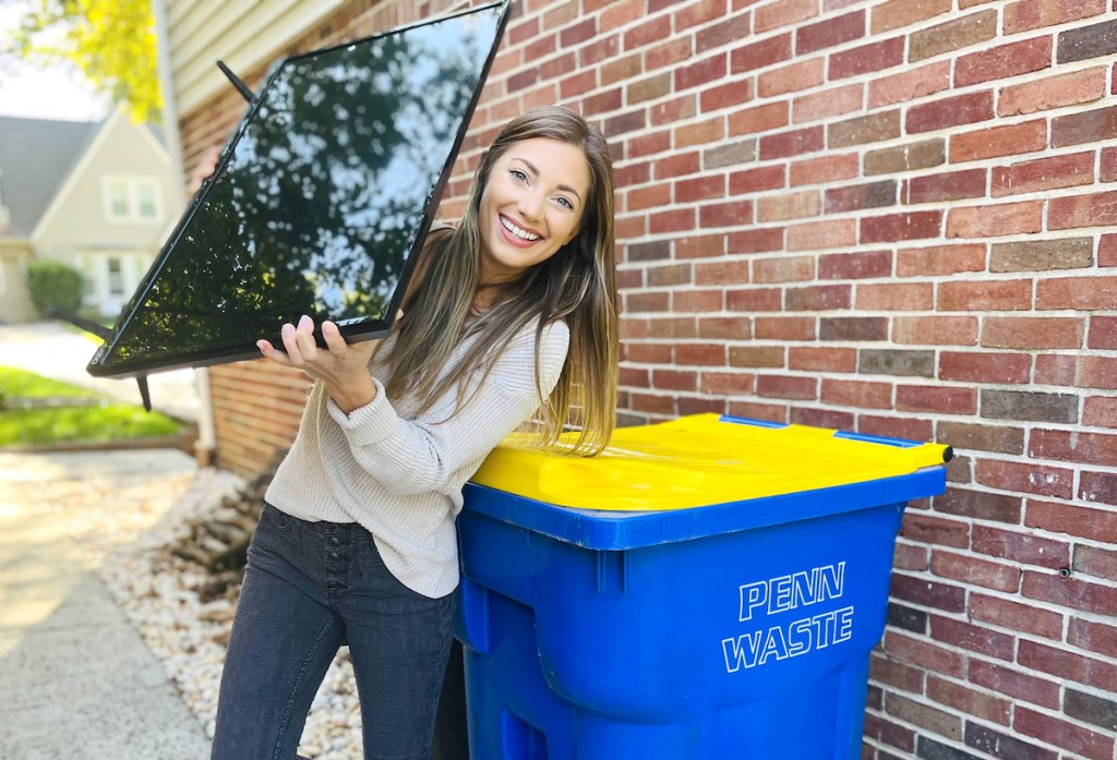 woman holding flat screen tv standing next to recycle bin