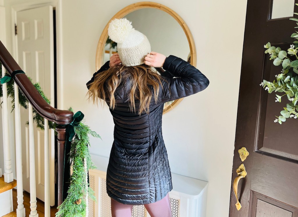 woman with black parka jacket and putting on winter hat in front of foyer mirror