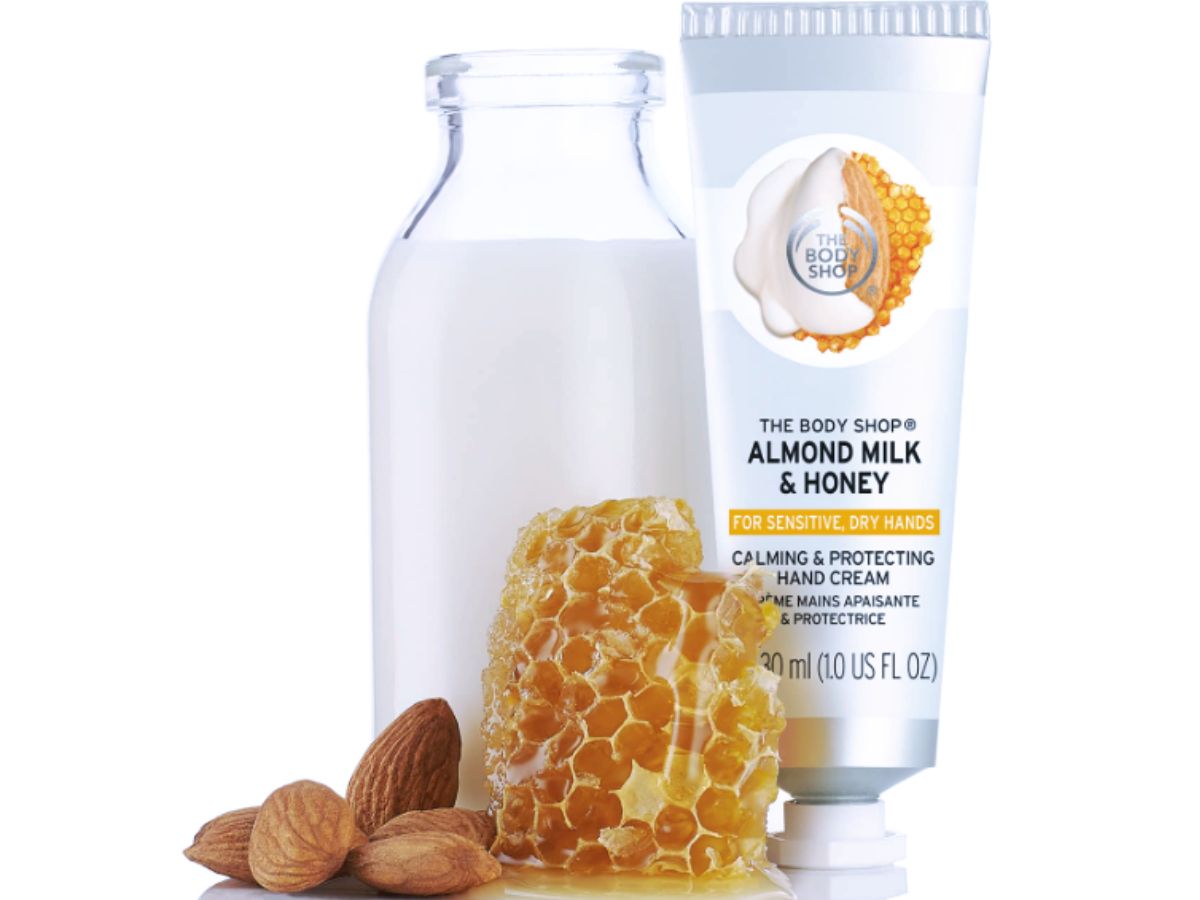 A tube of milk and honey hand cream pictured with a carafe of fresh milk, honey comb and almonds