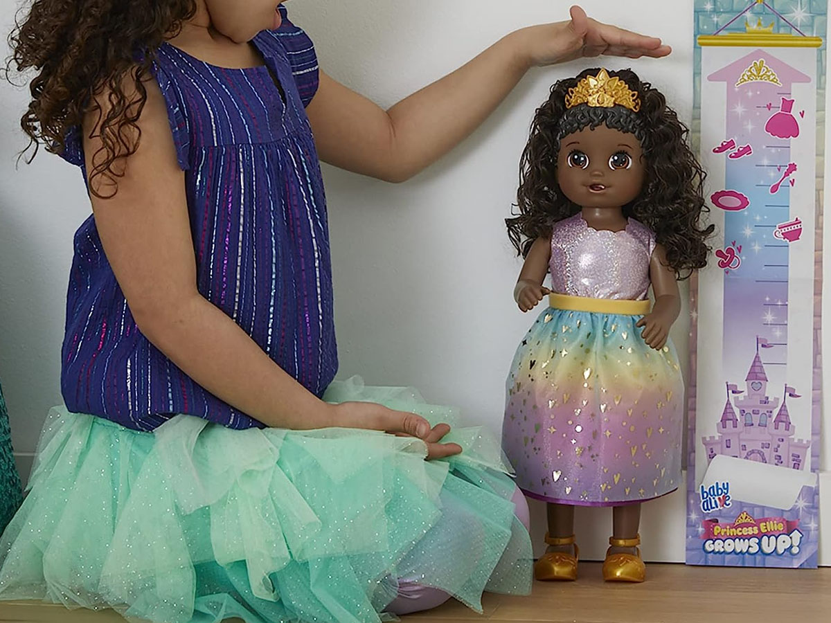 little girl measuring height of baby alive doll in rainbow dress and crown
