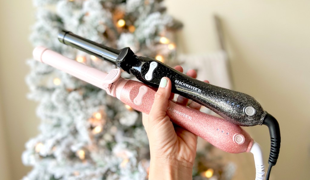 hand holding pink and black glitter beachwaver curling wands in front of christmas tree