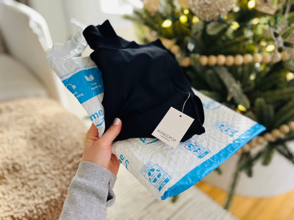 hand holding an amazon envelope bag and black mangopop bodysuit in front of christmas tree
