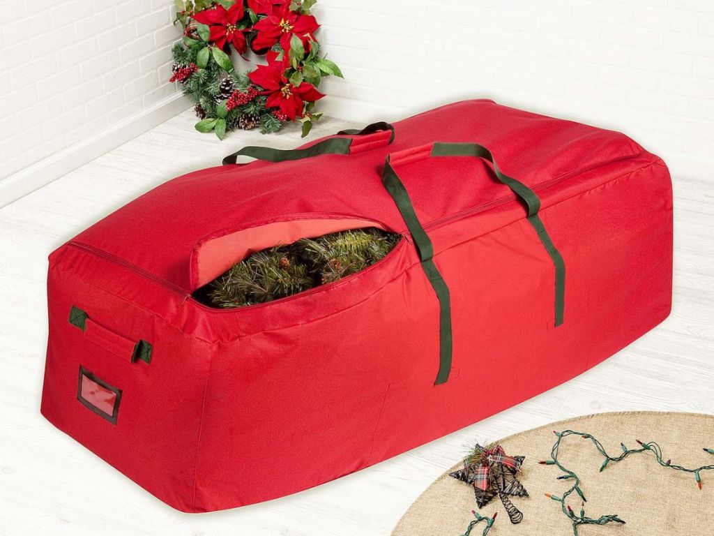 Honey-Can-Do Extra Large Christmas Tree Storage Bag with Wheels with tree inside sitting on floor with wreath behind it and lights beside it
