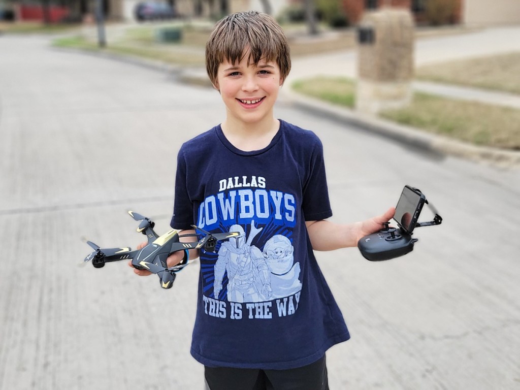 boy standing outside holding a drone in one hand and it's remote in the other