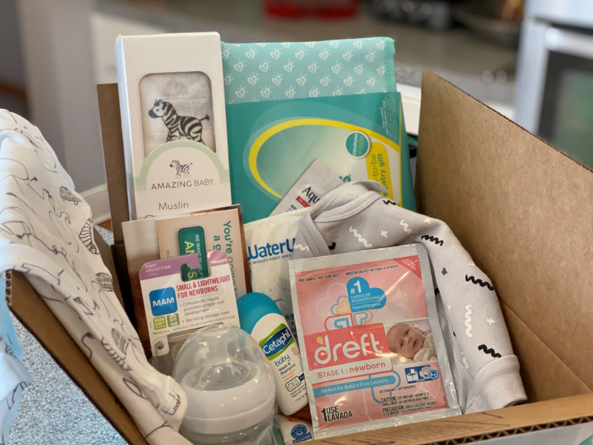 Box filled with baby items including Dreft sample, baby bottle, baby blanket, onesie, and Cetaphil sample.