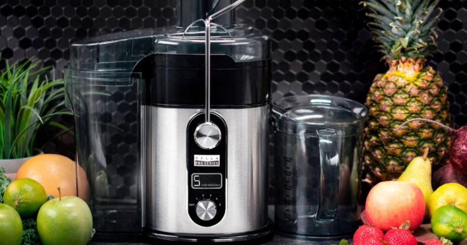 A stainless steel Bella Pro Series juice extractor surrounded by fresh fruit like apples, limes, a pear, and a pineapple on a countertop.