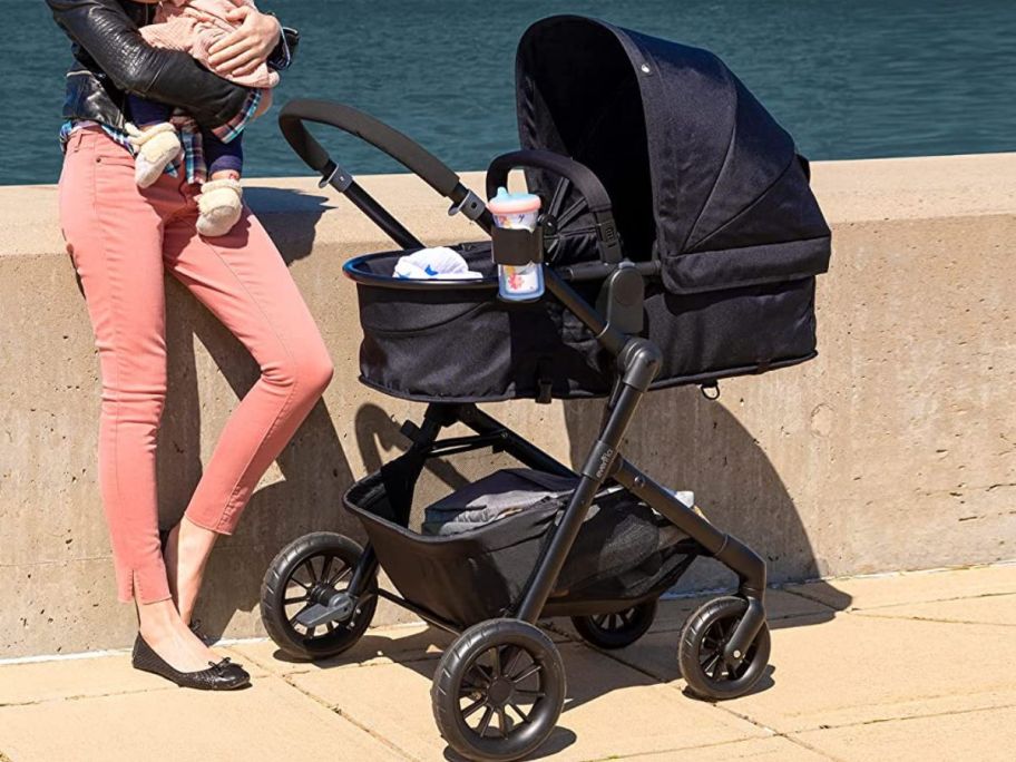 Woman and a baby standing next to the Evenflo Pivot travel system in carriage mode