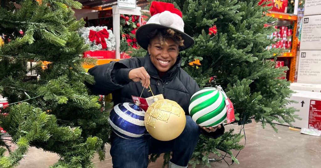 A lady in a hat holding 3 extra large christmas tree ornaments crouching down between 2 christmas trees