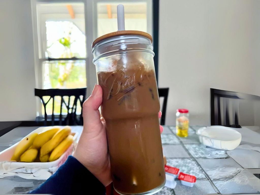 hand holding a tumbler style mason jar glass drinking glass with bamboo lid and straw, kitchen table with food in the background