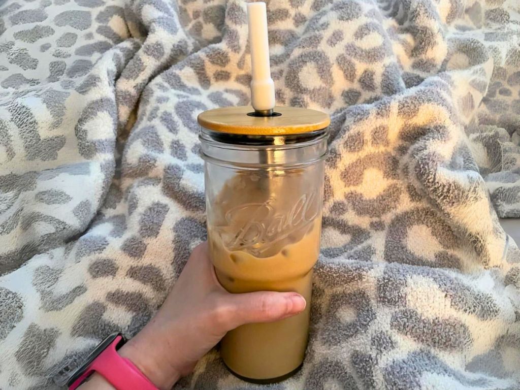 hand holding a tumbler style mason jar glass drinking glass with bamboo lid and straw, with a cheetah print blanket behind it