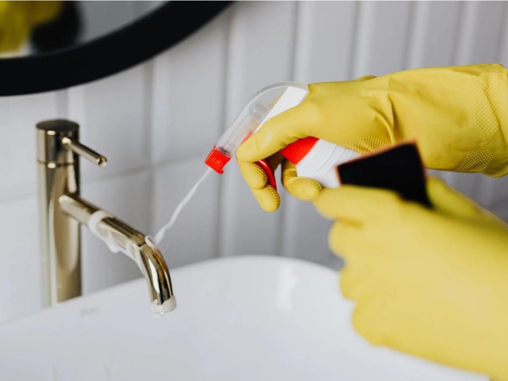gloved hands using cleaner spray from a bottle to clean a sink faucet
