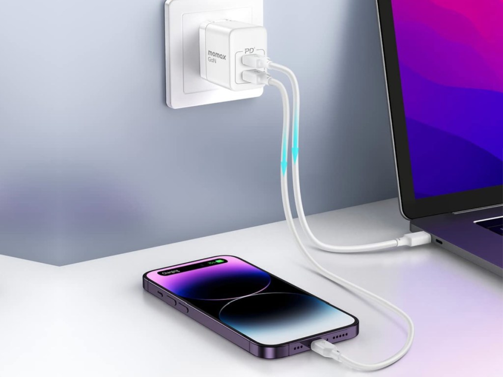 phone laying on a flat surface next to a laptop, both plugged into a wall charger