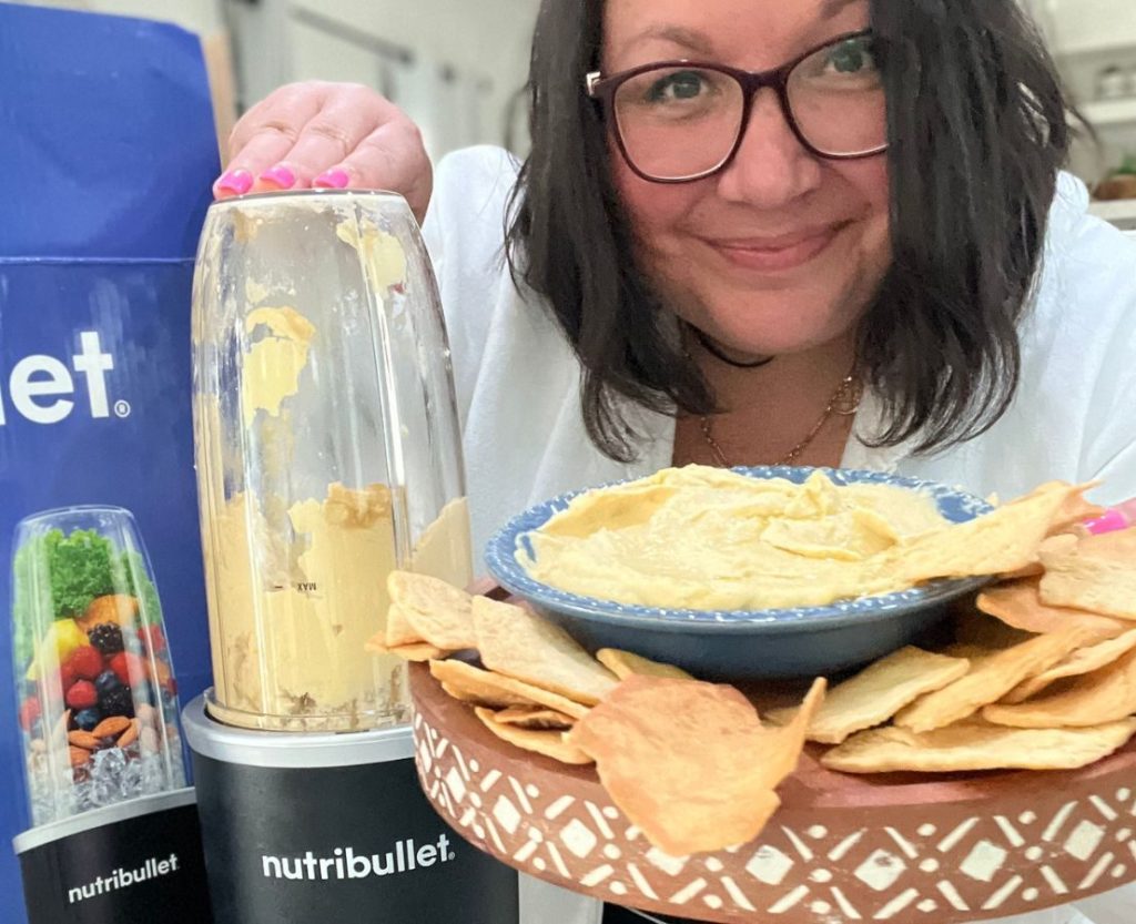 woman holding a plate of hummus and pita chips with her hand on a Nutribullet Pro+ Blender filled with hummus