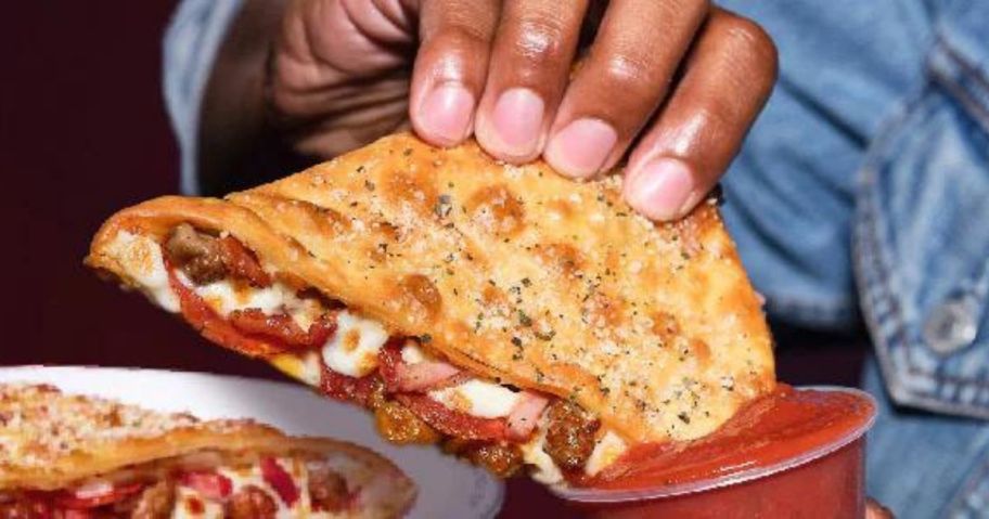 a hand dipping half of a pizza hut melt into a cup of marinara
