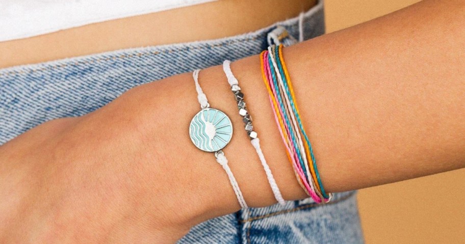 woman's arm wearing a stack of Pura Vida bracelets in various colors, some with beads, one with a sunshine charm