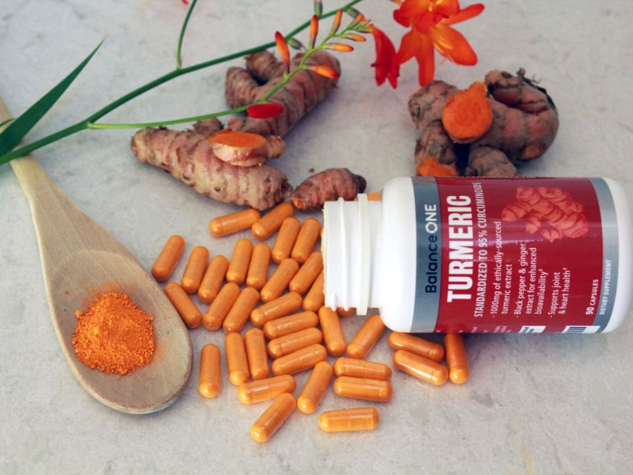 An open bottle of Balance One Turmeric with many capsules laid out next to a spoon of tumeric, whole tumeric roots and flowers
